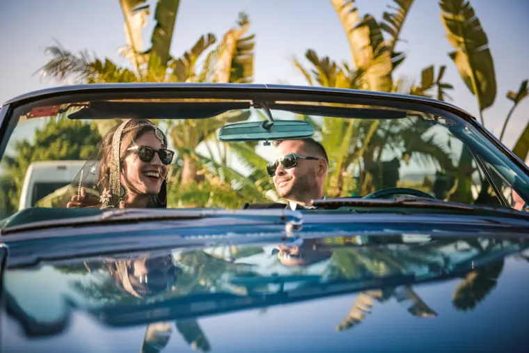 Couple in sunglasses laughing in a blue vintage convertible under palm trees