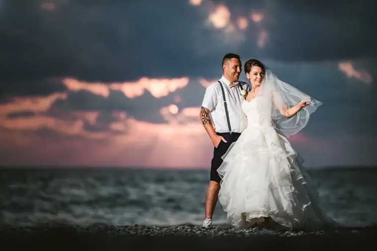 Groom in braces holding his bride on a pebble beach under a stormy pink sky