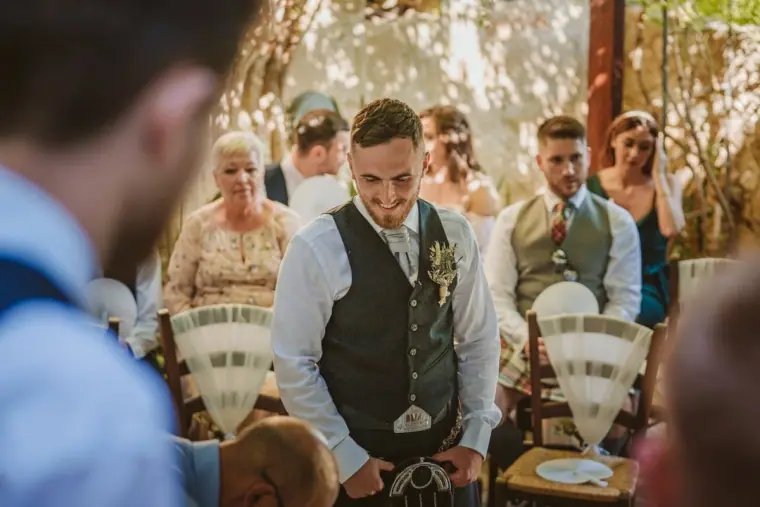Groom in a waistcoat and kilt grinning as he waits with guests seated behind
