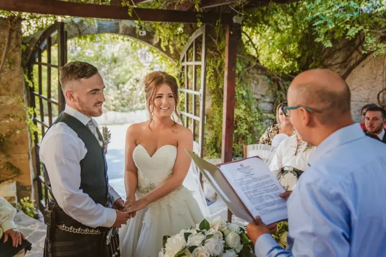 Hannah and Keith holding hands before the celebrant beneath a leafy stone archway