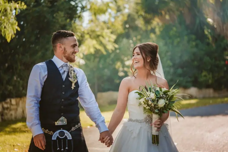 Newlyweds walking hand in hand along a sunny garden path, Hannah holding her bouquet
