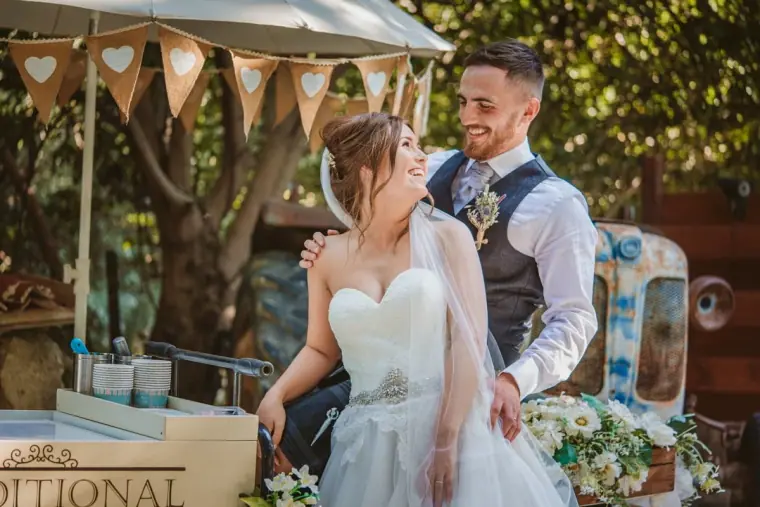 Bride and groom laughing together beside a rustic ice-cream cart and bunting