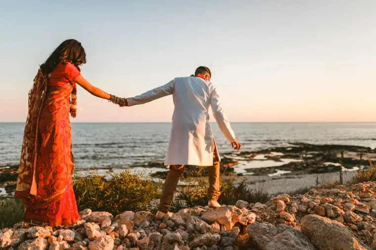 Raya in a red sari leading Adhib across the rocky shoreline at sunset