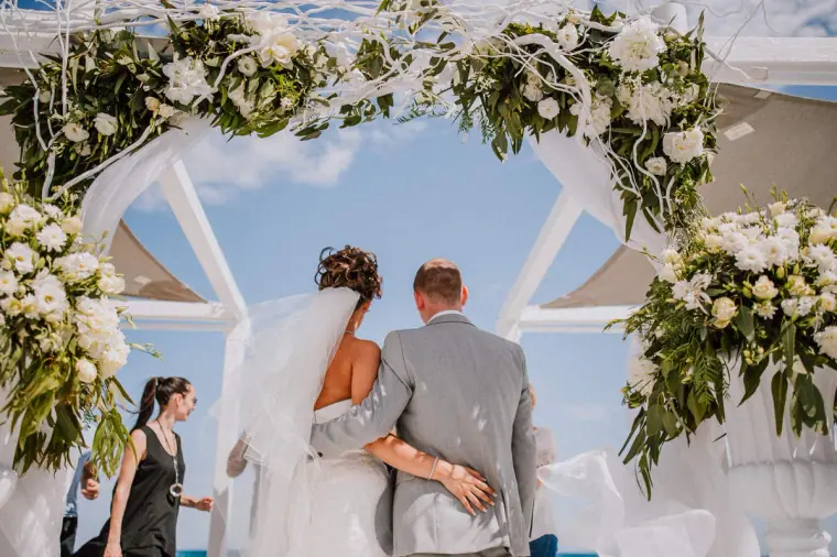 Bride and groom seen from behind under a white floral arch facing the sea at Galu Seaside, Larnaca