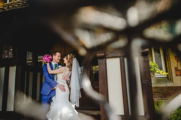 Groom in a blue suit embracing his bride, framed by a blurred iron gate at Cantley House, Berkshire