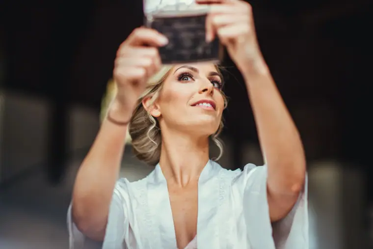 Bride in a satin robe taking a selfie on her phone while getting ready