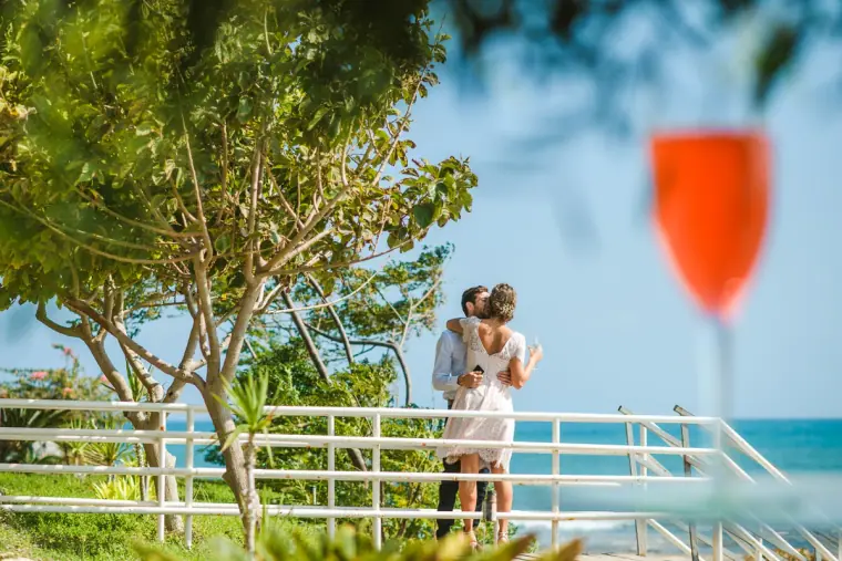 Rachel and Chris embracing by a white railing above the sea, a glass of rosé in the foreground, Pissouri