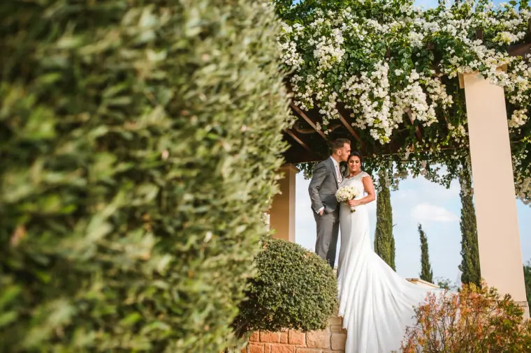 Anjali and Jonathan beneath a white-flowered pergola by a clipped hedge, Aphrodite Hills