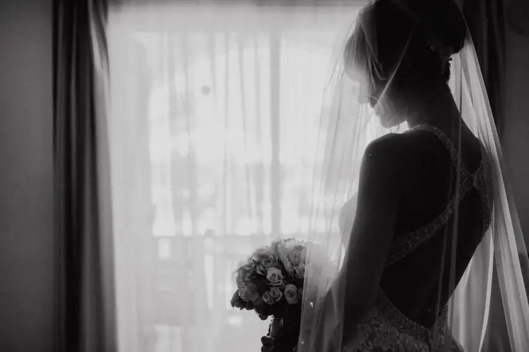 Black-and-white silhouette of a bride in her veil holding her bouquet by a curtained window