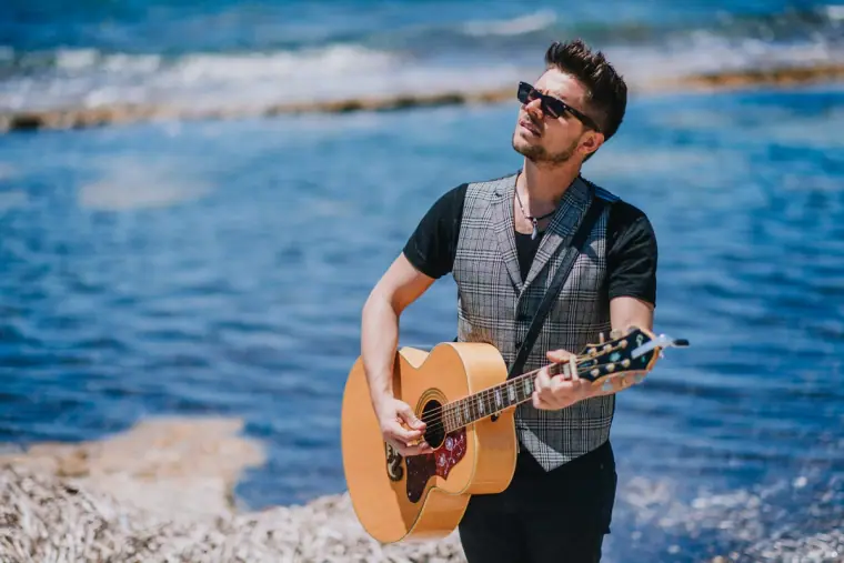 Guitarist in sunglasses and a waistcoat playing on the shore beside a blue sea