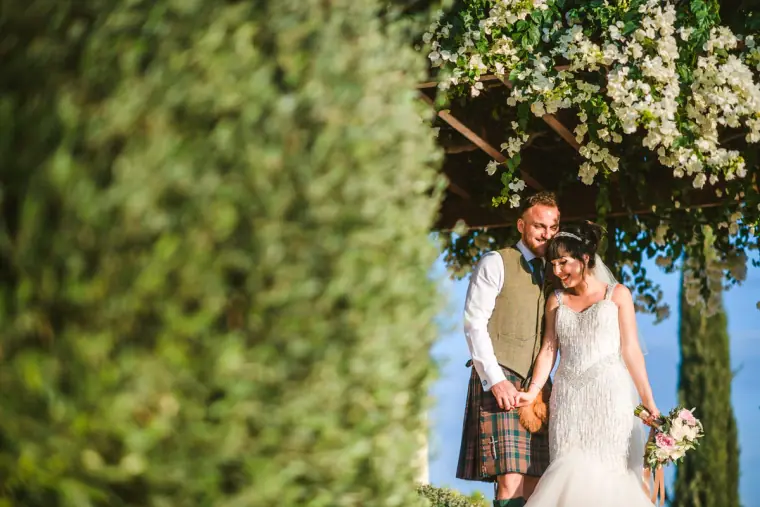 Marc in a kilt with Melissa under a white-blossom arch, framed by a hedge, Aphrodite Hills