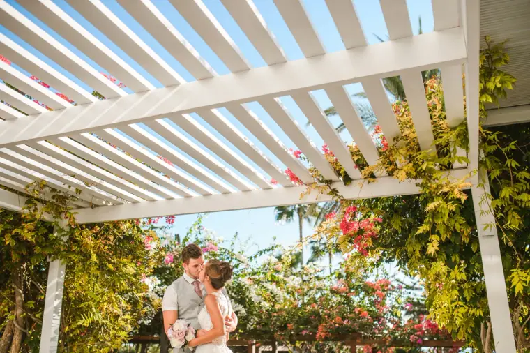 Naomi and Dale kissing under a white pergola draped with bougainvillea, Nissi Beach Resort