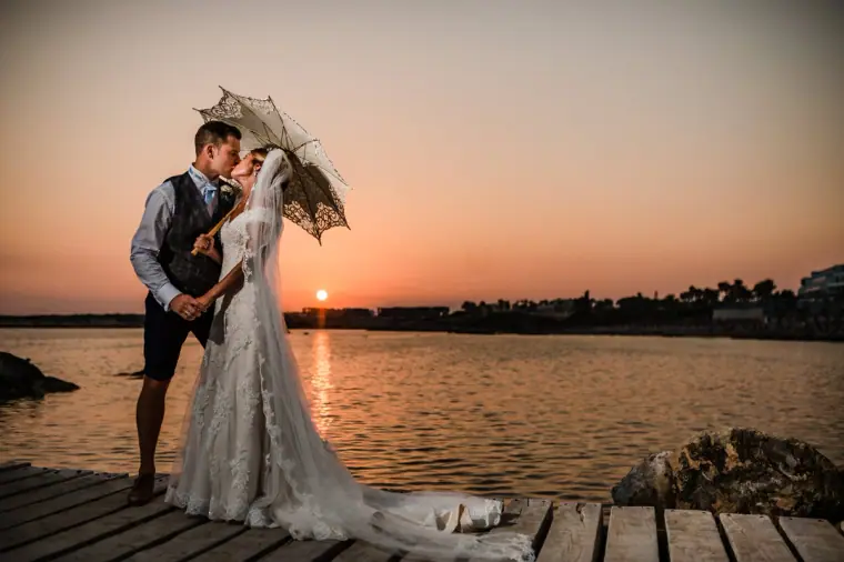 Chantelle and Paul kissing on a jetty at sunset under a lace parasol, Coral Beach Hotel