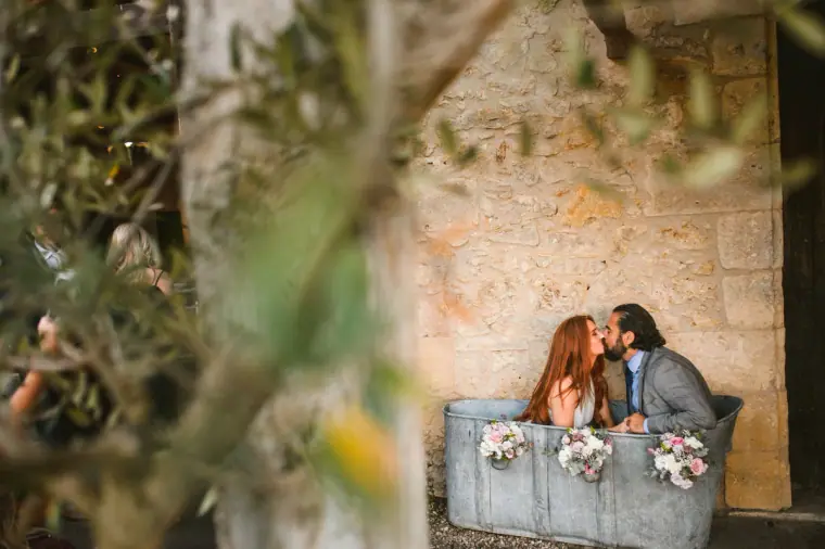 Sarah and Joe kissing behind a flower-filled stone trough, framed by olive leaves, Chateau Rigaud