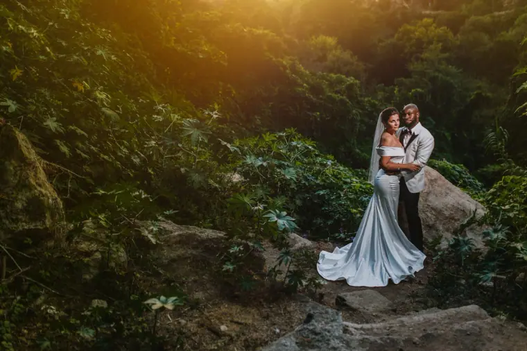 Reginald and Susannah embracing on rocks among lush greenery in golden light, Paphos