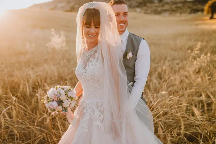 Bride with a sheer veil smiling in a golden field with the groom behind, Vasilias Nikoklis Inn