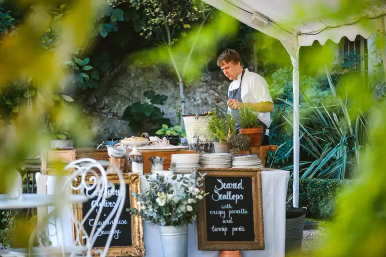 Chef at an outdoor food station with a chalkboard menu, seen through greenery, Chateau Rigaud
