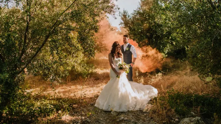 Bride and groom among olive trees as orange smoke drifts behind them, Vasilias Nikoklis Inn