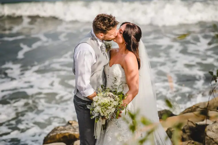 Couple kissing by crashing waves, bride holding a white bouquet, Royal Apollonia, Limassol