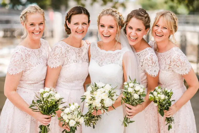 Bride laughing with bridesmaids in matching lace dresses holding white bouquets, Ayia Napa
