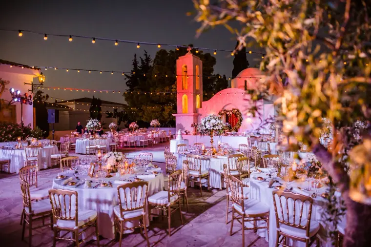 Reception tables under festoon lights lit in pink beside a small chapel at dusk, Anassa