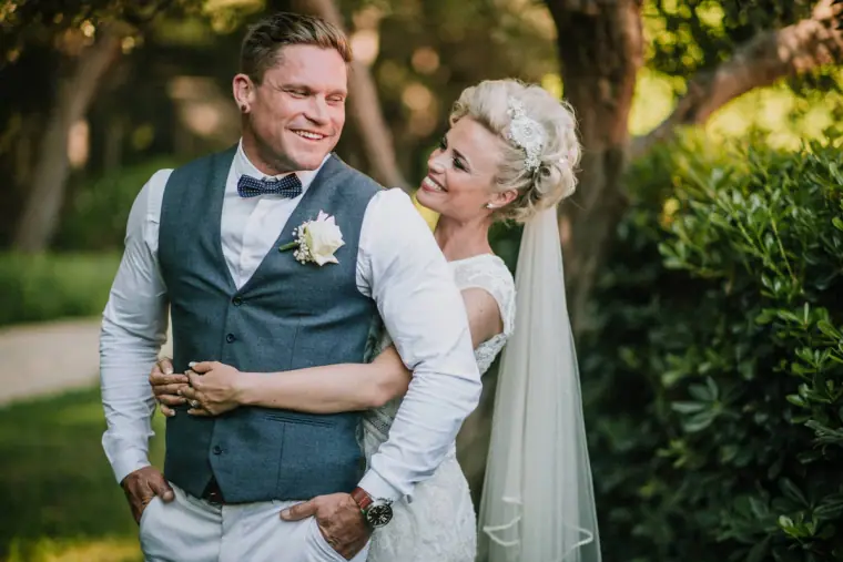 Groom in a blue waistcoat laughing as the bride hugs him from behind, Elysium, Paphos