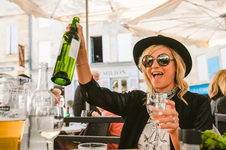 Laughing guest in a hat and sunglasses holding up a wine bottle at a café, Bordeaux