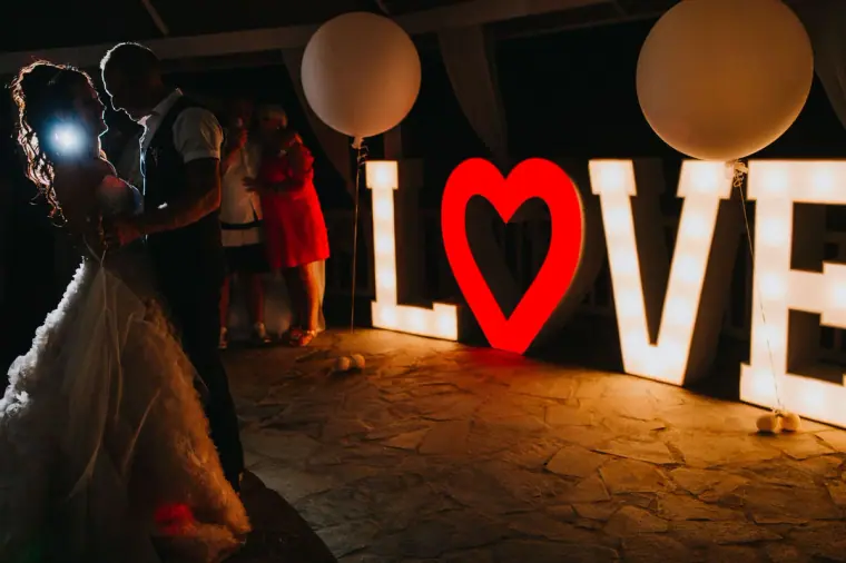 Sarah and Martin beside giant light-up LOVE letters with a red heart at night, Nissi Beach