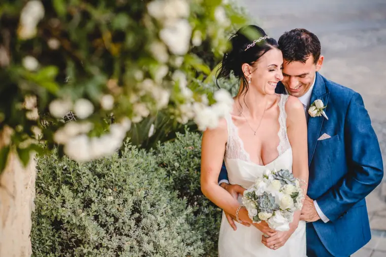 Groom in a blue suit cuddling his bride behind a white flowering shrub, Aphrodite Hills