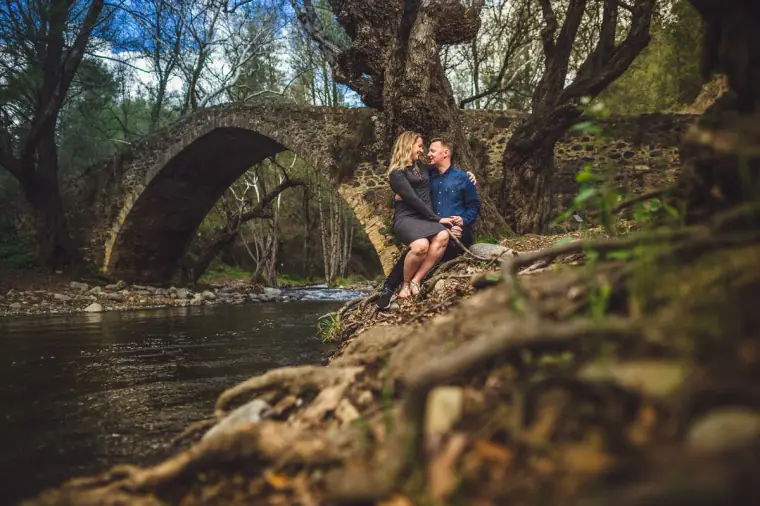 Couple sitting by a river beside an old stone arched bridge in the Troodos during an engagement shoot