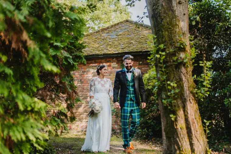 Helen and Ali walking out of a leafy garden by a brick cottage, groom in tartan trousers, Windsor