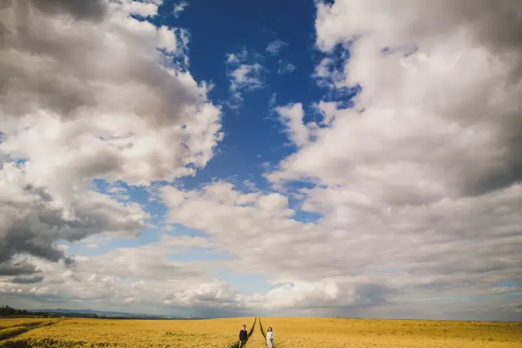 Tiny couple standing in a vast golden field beneath a huge cloudy sky in Scotland