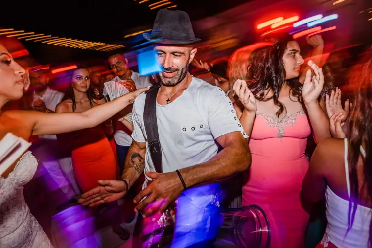 Guest in a fedora dancing among friends under colourful party lights, Anassa