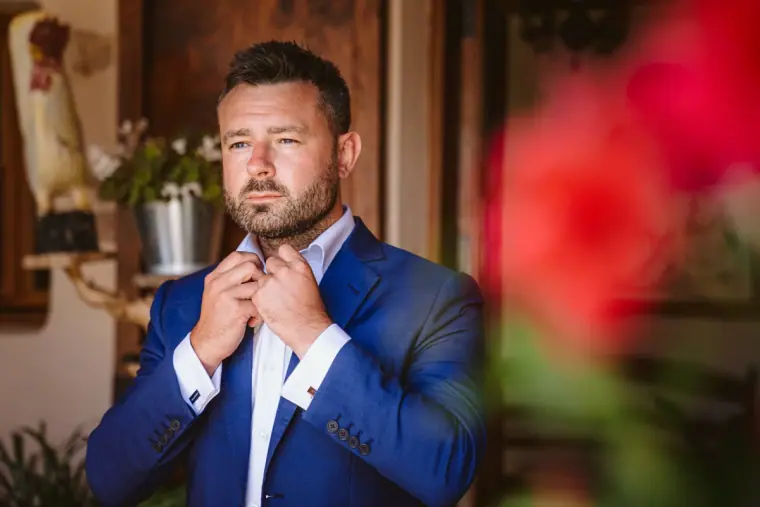 Iain in a blue suit fastening his collar beside a blurred red flower