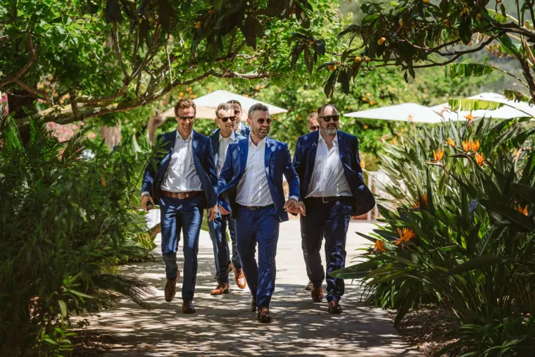 Groom and groomsmen in navy suits striding along a shady garden path