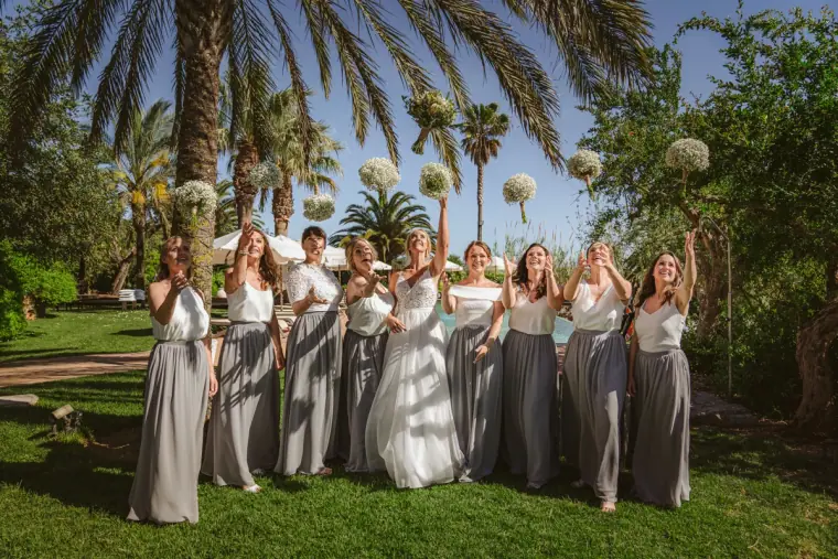 Bridesmaids in grey maxi skirts cheering with raised arms on a palm-lined lawn