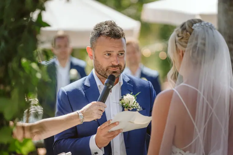 Iain reading his vows into a microphone as Sophie listens under a veil