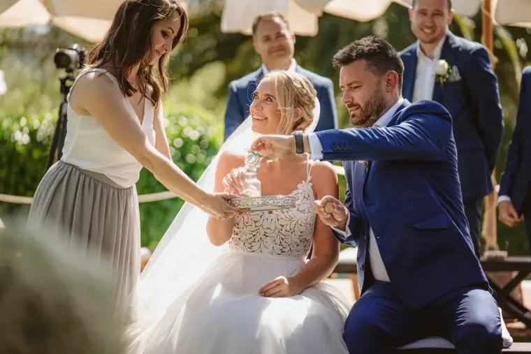 Sophie and Iain pouring into a glass vessel during a unity ceremony as a bridesmaid holds a tray