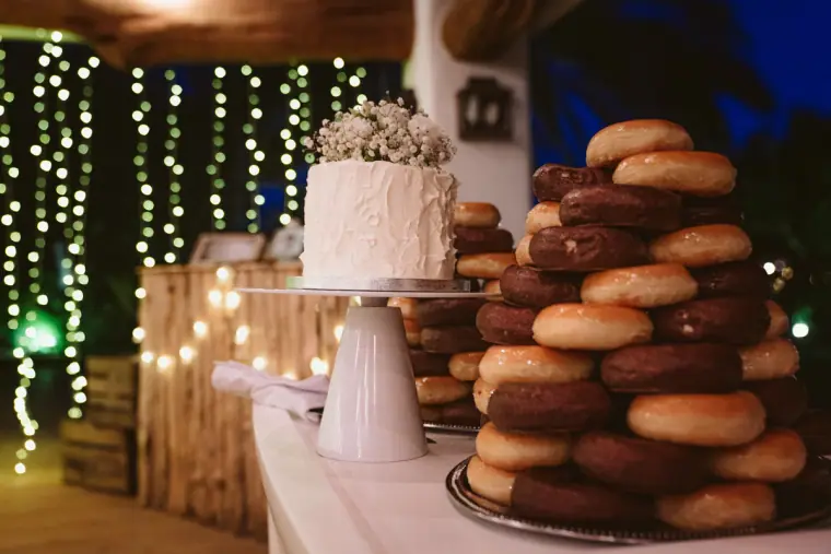 Wedding cake beside a tower of doughnuts in front of curtains of fairy lights