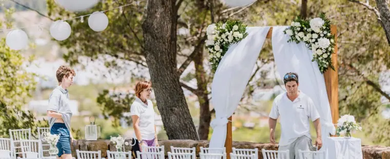 Team preparing a garden ceremony with a floral, fabric-draped arch under trees at Can Curreu, Ibiza