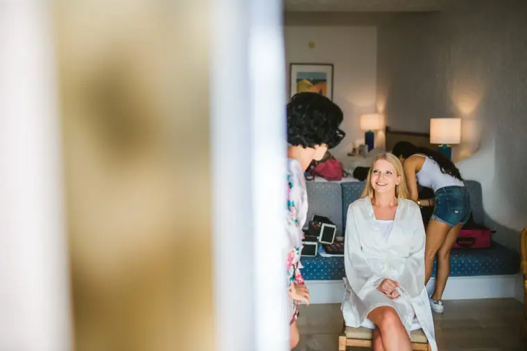 Charlotte sitting in her white robe while bridesmaids get ready in the hotel room behind her