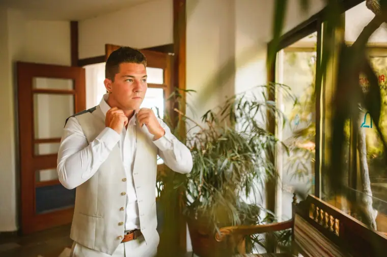 Nathan fastening his shirt collar in a light, plant-filled hotel room