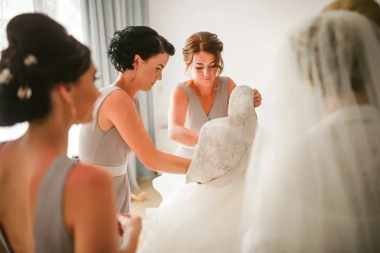 Bridesmaids in grey dresses lifting the bride's lace gown