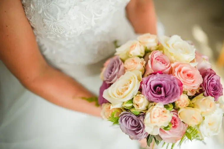 Close-up of a pink, purple and cream rose bouquet held against a lace wedding dress