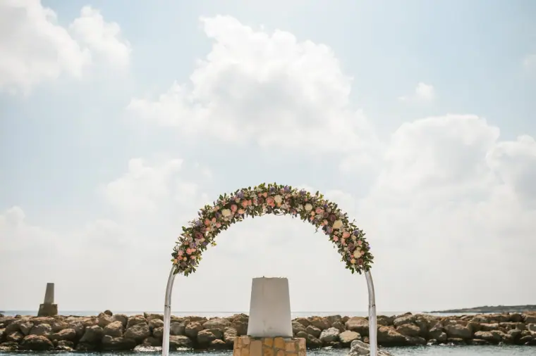 Floral wedding arch on the stone jetty at Coral Beach Hotel against a cloudy sky