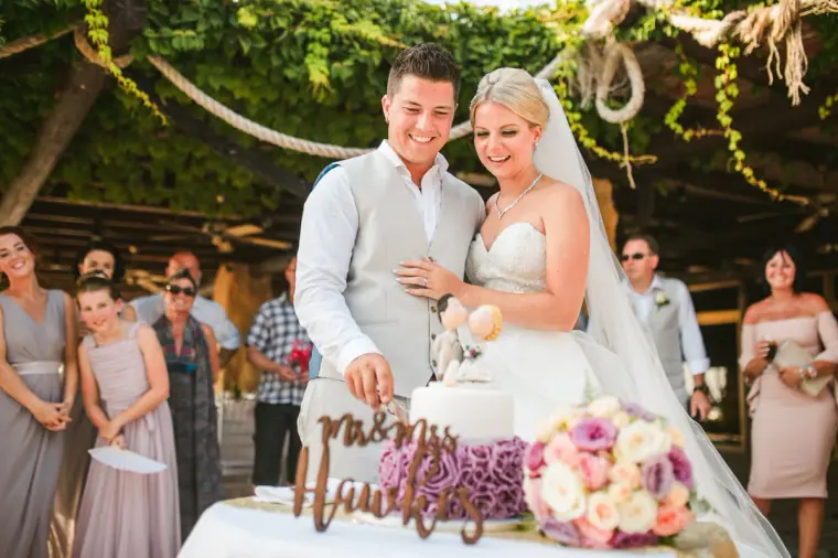 Charlotte and Nathan cutting their wedding cake as guests watch under the pergola