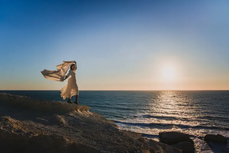 Aiko's dress billowing in the wind on a cliff edge as the sun sets over the sea