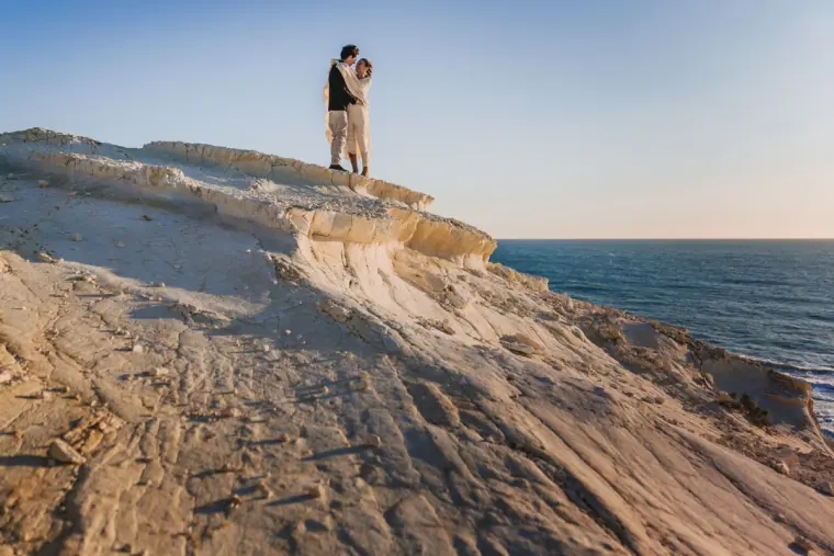 Aiko and Erez embracing on top of a pale limestone cliff above the sea