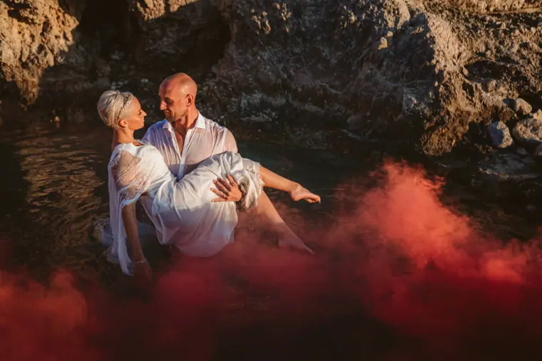 Luke carrying Alana through the water as red smoke drifts across the rock pool