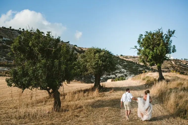 Amie and Graeme walking hand in hand through a dry field dotted with carob trees